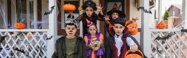 Meninas mostrando gestos assustadores perto de amigos multiétnicos em trajes de Halloween, banner — Fotografia de Stock