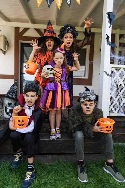 Multiethnische Kinder in Halloween-Kostümen halten Tricks in der Hand oder behandeln Eimer, während sie grimmig sind und in der Nähe des Hauses gruselige Gesten zeigen — Stockfoto