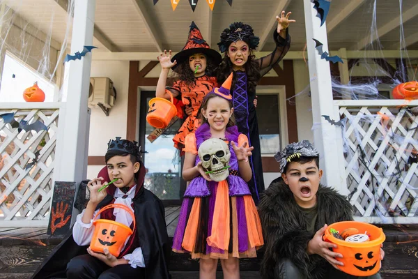 Amis multiethniques en costumes d'Halloween tenant des seaux de bonbons et grimacer près de chalet décoré — Photo de stock