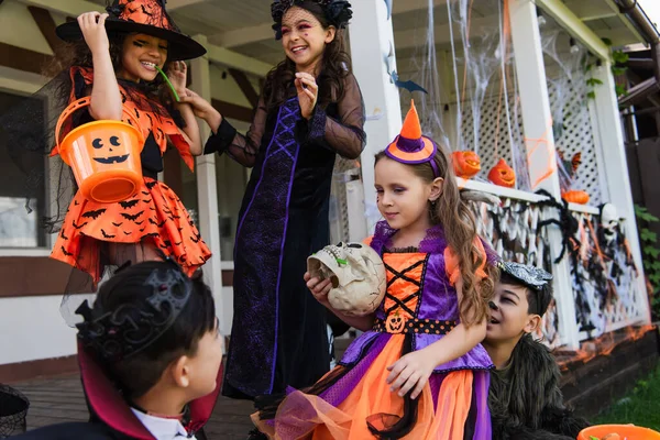 Девушка в костюме клоуна держит череп рядом с весёлыми мультикультурными друзьями и украшенным коттеджем — стоковое фото