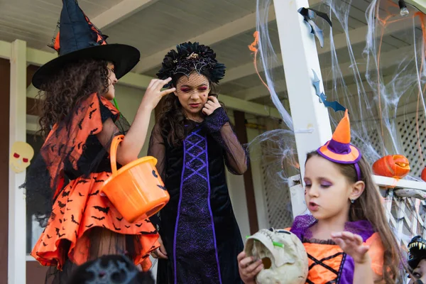 Девушки в костюмах на Хэллоуин держат череп и трюк или корзину для выпечки возле украшенного коттеджа — стоковое фото