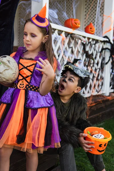 Boy in werewolf costume holding bucket with candies and grimacing near girl with skull — Stock Photo
