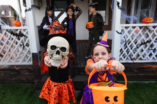 Chicas en disfraces de Halloween sosteniendo cráneo y truco o tratar cubo cerca de casa y borrosa interracial amigos - foto de stock