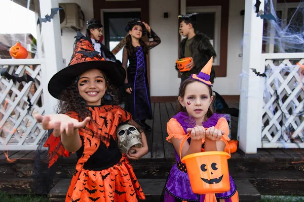 Lächelndes Mädchen im Hexenkostüm mit ausgestreckter Hand neben Freundin mit Halloween-Eimer — Stockfoto