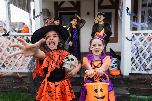Girl in witch costume screaming with outstretched hand near smiling friend with trick or treat bucket — Stock Photo