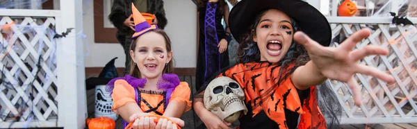 Girl in witch costume holding skull and grimacing with outstretched hand near friend, banner — Stock Photo