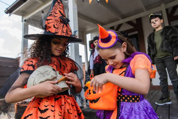 Menina em bruxa fantasia alimentando crânio com biscoito perto amigo olhando em balde halloween — Fotografia de Stock