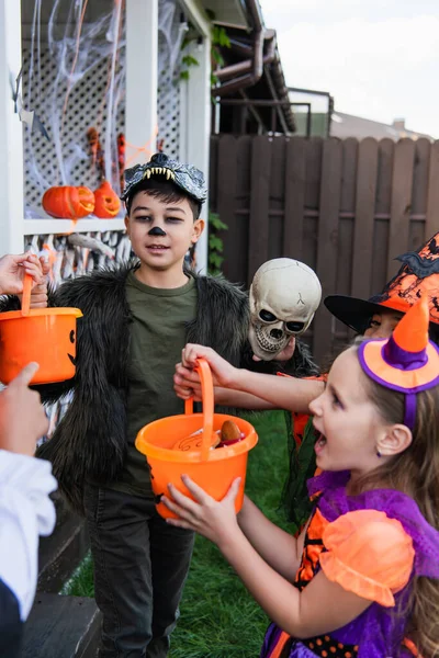 Asiático chico en halloween traje celebración cubo con caramelos cerca amigo en patio trasero - foto de stock