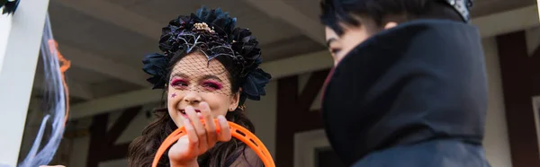 Smiling girl in halloween costume looking at blurred asian friend outdoors, banner — Stock Photo