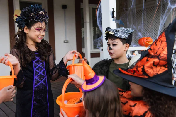 Smiling kid holding buckets near interracial friends in halloween costumes outdoors — Stock Photo