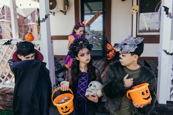 Preteen girl holding bucket with candies near asian friends in halloween costumes outdoors — Stock Photo