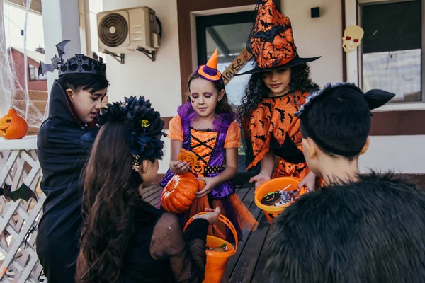 Interrassische Kinder in Halloween-Kostümen und Schminke halten Eimer mit Bonbons in der Nähe des Hauses — Stockfoto