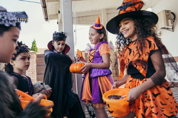 Alegre niño sosteniendo calabaza y galletas cerca de amigos interracial en disfraces de Halloween en el patio trasero - foto de stock