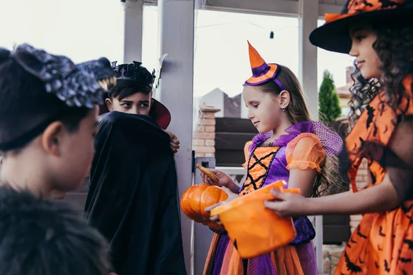 Niño sosteniendo calabaza y galleta cerca borrosa amigos multiétnicos en trajes de Halloween al aire libre - foto de stock