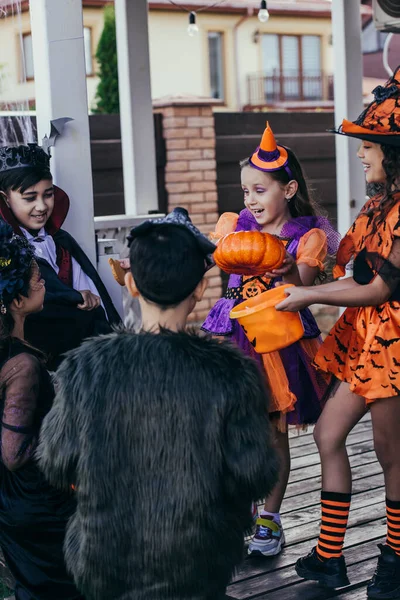 Crianças multiétnicas felizes segurando balde perto de amigos em trajes de Halloween ao ar livre — Fotografia de Stock