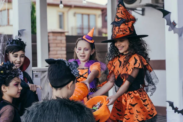 Niño sonriente en traje de Halloween sosteniendo cubo con caramelos cerca de amigos interracial al aire libre - foto de stock