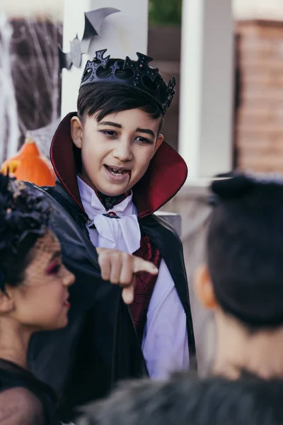 Asiático chico en halloween vampiro traje apuntando con el dedo a borrosa amigos al aire libre - foto de stock