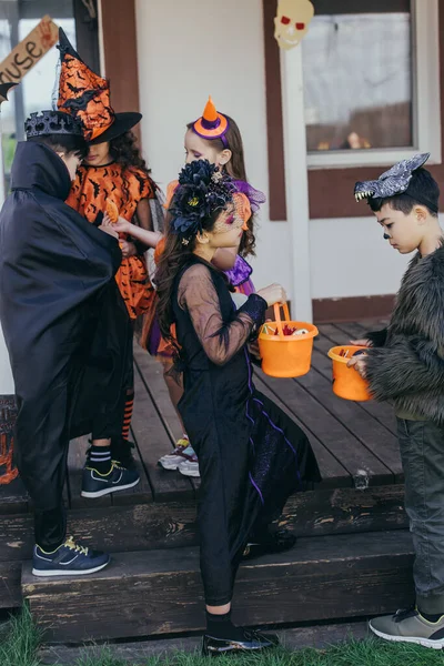 Multiethnic kids holding buckets with candies near friends in halloween costumes outdoors — Stock Photo