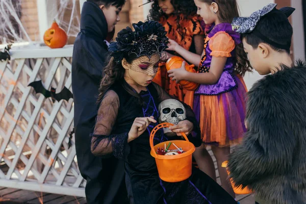 Shocked chica en traje celebración cubo con caramelos cerca asiático amigo durante la celebración de Halloween al aire libre - foto de stock