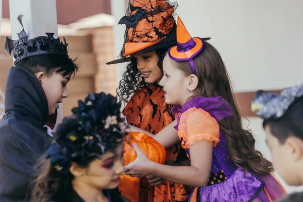 Sonriente chica en halloween traje celebración calabaza cerca interracial amigos al aire libre - foto de stock