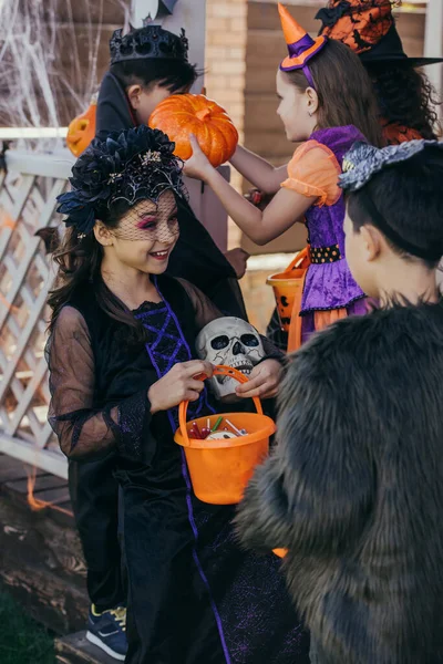 Alegre niño en halloween traje celebración cráneo y cubo con caramelos cerca asiático amigo al aire libre — Stock Photo