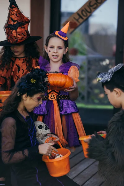 Chica preadolescente sosteniendo calabaza de Halloween cerca de niños multiétnicos con cubos al aire libre - foto de stock
