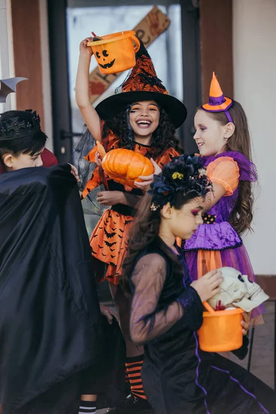 Menina alegre em chapéu de bruxa segurando balde perto de amigos multiétnicos durante a celebração halloween ao ar livre — Fotografia de Stock