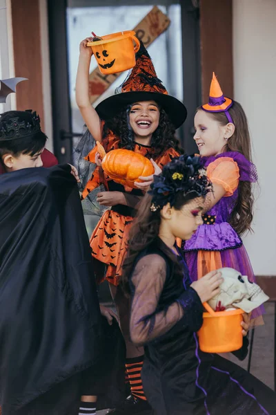 Chica alegre en sombrero de bruja sosteniendo cubo cerca de amigos multiétnicos durante la celebración de Halloween al aire libre - foto de stock