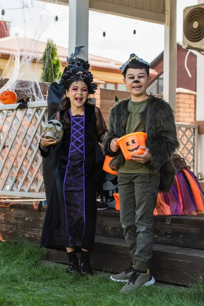 Positivo interracial niños en halloween trajes celebración cráneo y cubo en hierba en patio trasero - foto de stock