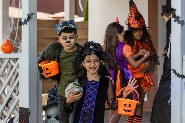 Happy girl in halloween costume holding skull near asian friend with bucket and kids in backyard — Stock Photo