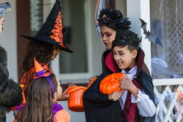 Sonriente asiático chico con maquillaje apuntando a calabaza cerca de amigos en halloween trajes en patio trasero - foto de stock