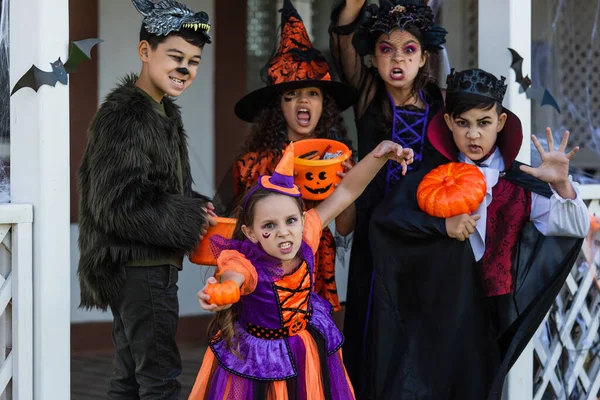 Мультиэтнические дети в костюмах на Хэллоуин гримасничают перед камерой возле дома на открытом воздухе — стоковое фото