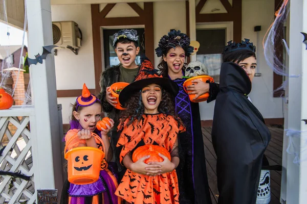 Excited multiethnic children holding buckets with candies near decor during halloween celebration in backyard — Stock Photo