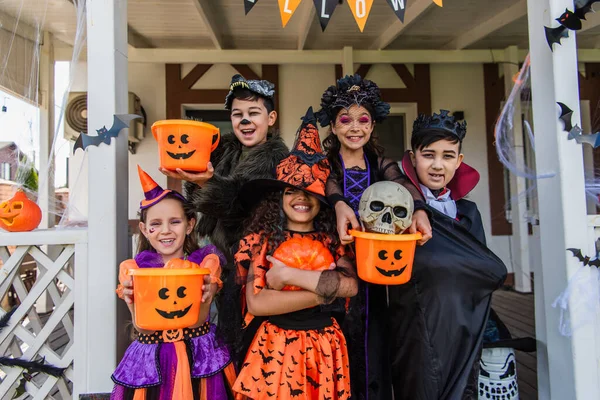 Niños multiétnicos felices en trajes de Halloween sosteniendo cubos y calabaza cerca de la casa al aire libre - foto de stock