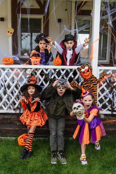 Enfants multiethniques en costumes d'Halloween grimacant près de Jack o lanternes dans la cour — Photo de stock