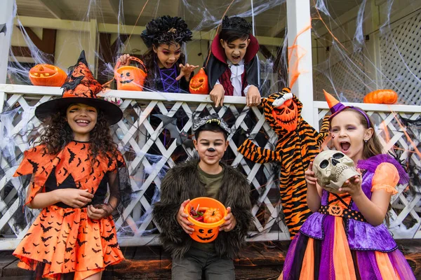 Menino asiático em traje de Halloween segurando balde com doces perto de amigos e decoração no quintal — Fotografia de Stock