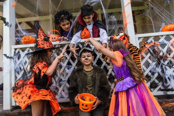 Дети-подростки в костюмах на Хэллоуин гримасуют рядом с азиатским другом с ведром на открытом воздухе — стоковое фото
