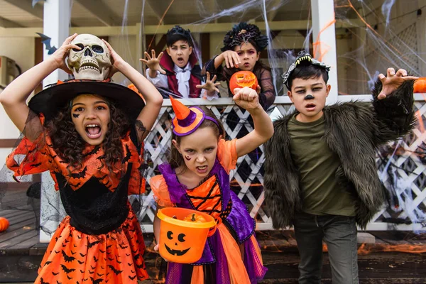 Preteen multiethnic kids with bucket and skull grimacing at camera near friends and house with halloween decor outdoors — Stock Photo