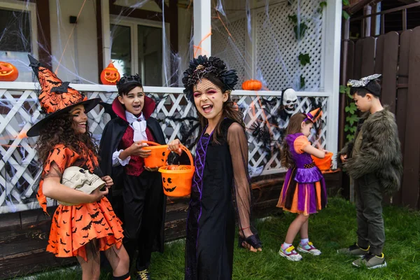 Fröhliches Mädchen hält Halloween-Eimer in der Nähe von interrassischen Freunden mit Totenkopf im Freien — Stockfoto