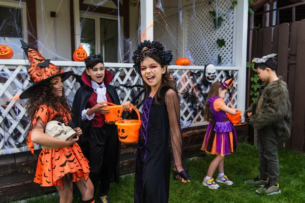 Cheerful girl holding halloween bucket near interracial friends with skull outdoors — Stock Photo