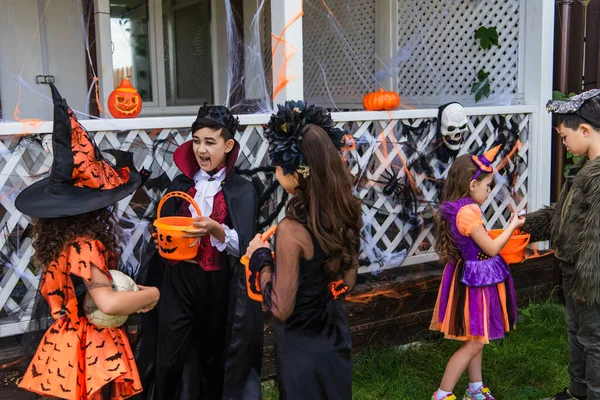 Crianças multiétnicas em trajes de Halloween segurando baldes perto da teia de aranha e decoração na casa — Fotografia de Stock