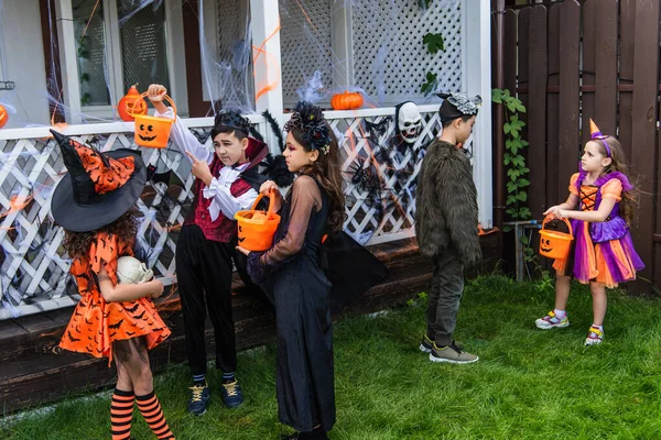 Asian boy in halloween costume pointing at bucket near girls in backyard — Stock Photo