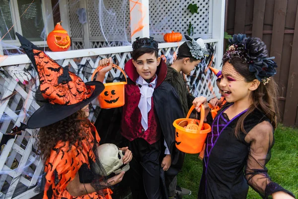 Asiático chico en traje mirando cámara cerca alegre amigos con cubo hablando durante halloween celebración al aire libre - foto de stock