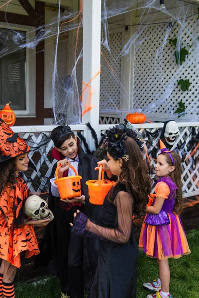 Sonriendo niños preadolescentes en trajes de Halloween mirando cubos con caramelos cerca de amigos interracial al aire libre - foto de stock