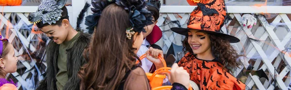 Chica preadolescente sosteniendo cubo cerca de amigos interracial en trajes de Halloween al aire libre, pancarta - foto de stock