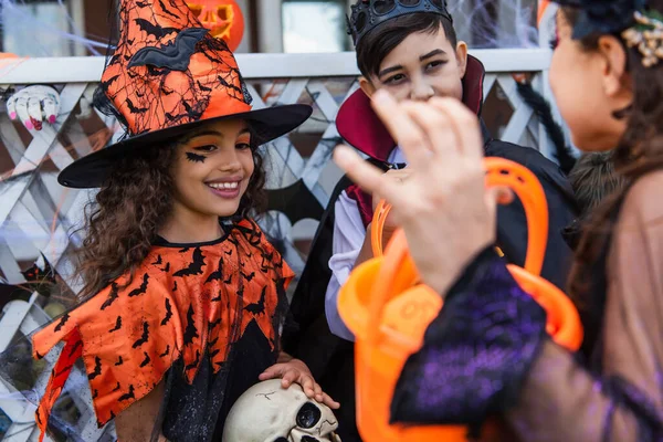 Menina sorridente em chapéu de bruxa segurando crânio perto de amigos multiétnicos com balde durante a festa de Halloween ao ar livre — Fotografia de Stock
