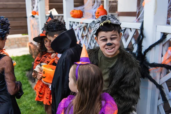 Asiático niño en halloween traje mueca en cámara cerca de amigos y decoración al aire libre - foto de stock