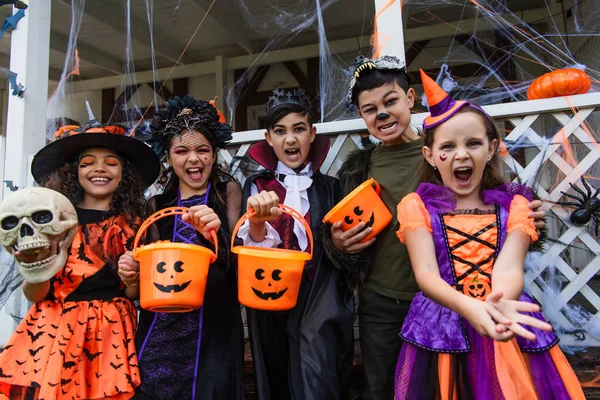 Smiling interracial kids grimacing and holding halloween buckets outdoors — Stock Photo