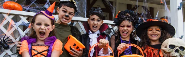 Niños multiétnicos sosteniendo cubos mientras celebran Halloween al aire libre, pancarta - foto de stock
