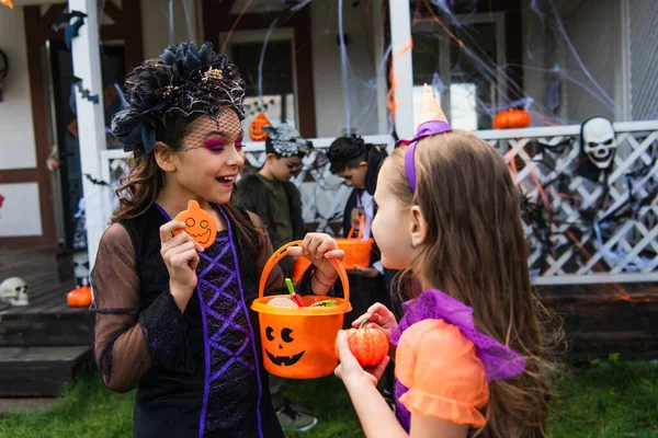 Улыбающаяся девушка держит ведро на Хэллоуин с конфетками рядом с другом с тыквой на заднем дворе — стоковое фото